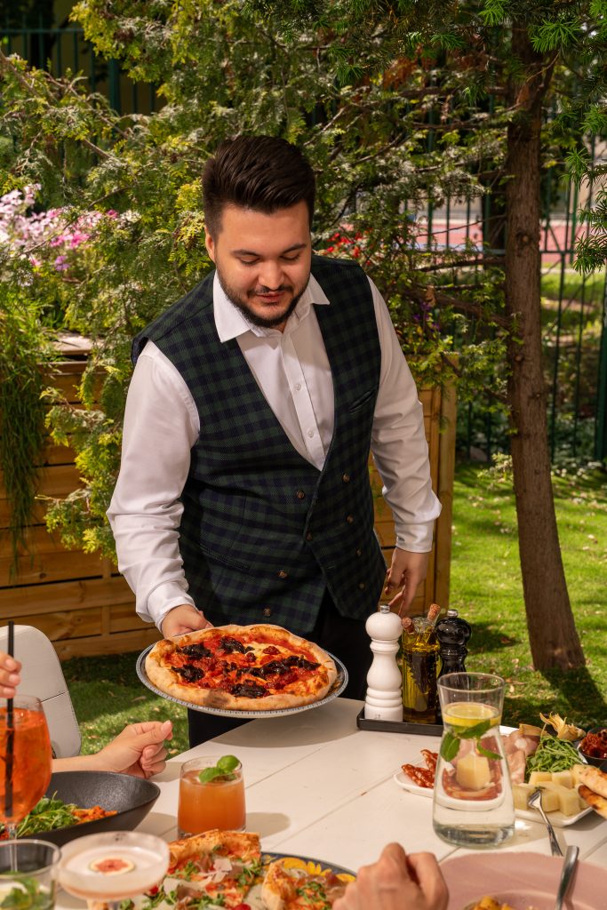 Uśmiechnięty kelner La Tropea podaje pizzę gościom w letnim ogródku restauracji